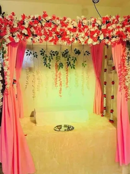 A lovely home mandap featuring a canopy of red and white flowers with flowing pink drapes. The backdrop includes hanging green vines, adding a touch of natural elegance to the festive setup.