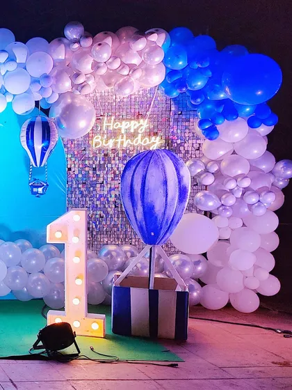 A dreamy hot air balloon theme for a first birthday. This magical setup features a large balloon basket prop, a shimmering sequin backdrop, and a cloud-like balloon garland in shades of blue and white. The light-up number '1' and neon 'Happy Birthday' sign add a warm glow.