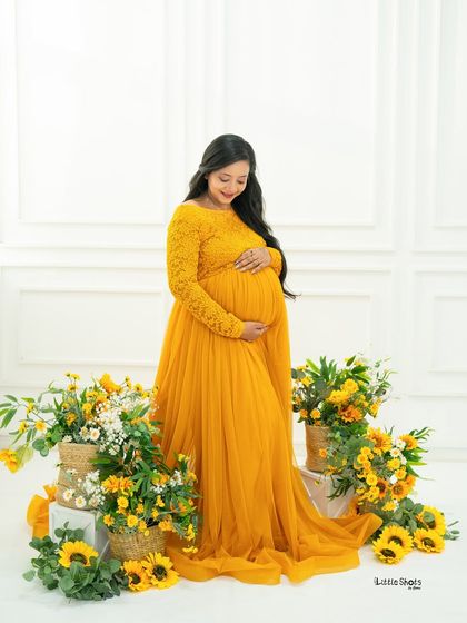 A cheerful portrait of an expecting mother in a flowing yellow gown. She looks down at her bump with a smile, surrounded by an arrangement of sunflowers and other yellow blooms.