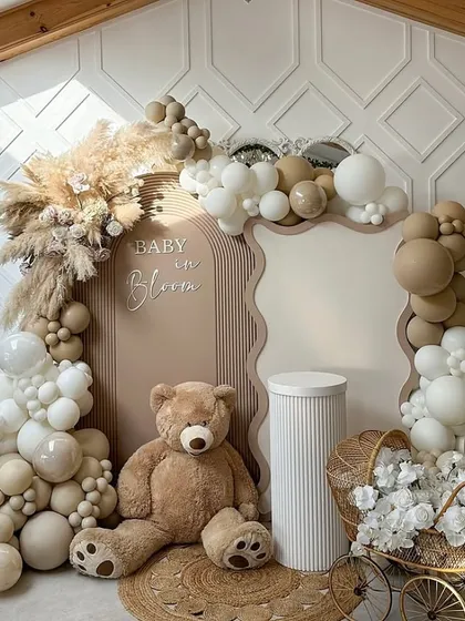 A "Baby in Bloom" themed baby shower backdrop. This setup features a large teddy bear, a beautiful balloon arch in neutral tones, pampas grass, and a vintage-style pram filled with white flowers for a boho-chic look.