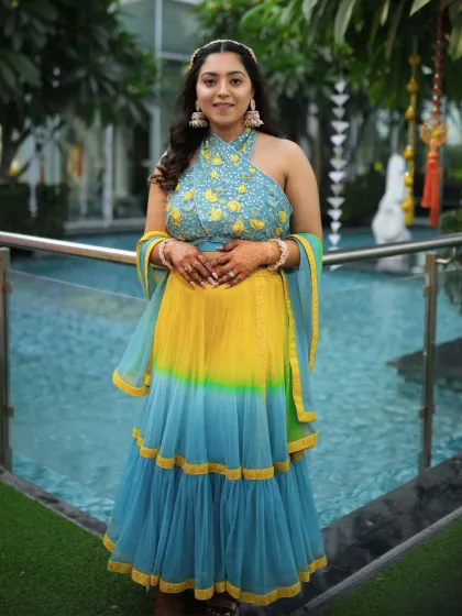 A full-length shot of the bride in her stunning yellow and teal Haldi lehenga. The makeup is soft and glamorous, with warm flushed cheeks and sparkling eyes.
