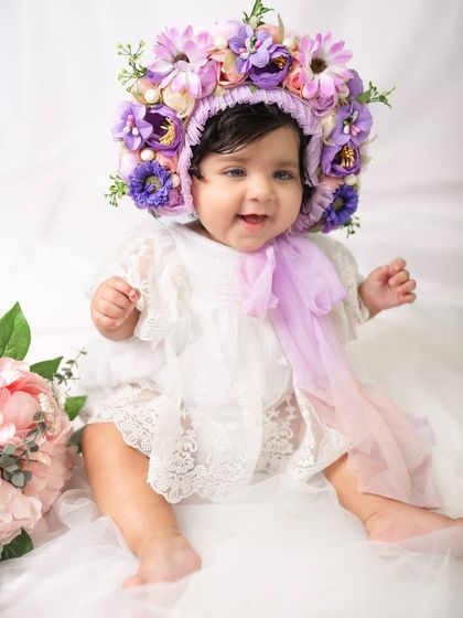 This happy eight-month-old is sitting up and smiling, surrounded by soft white fabric and wearing a gorgeous handmade floral bonnet.