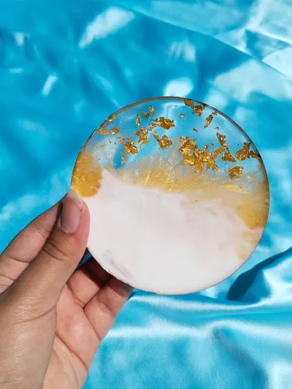 A round resin coaster featuring a beautiful ocean-inspired design. The piece blends white and translucent blue resin, with gold flakes scattered along the edge for a touch of luxury.