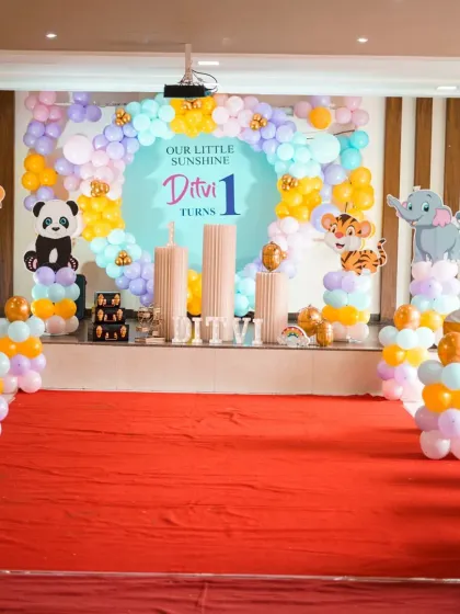A wide shot of the "Our Little Sunshine" first birthday stage. The setup includes a large backdrop, a pastel balloon arch, and cute animal cutouts like a panda, lion, and tiger on balloon pillars.