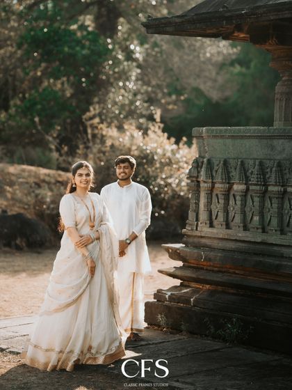 A full-length portrait of the couple standing proudly in their coordinated white ethnic outfits, set against the warm, sunlit stone of a heritage site.