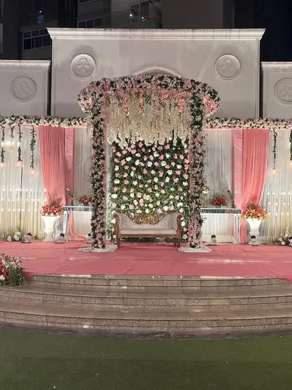 A beautiful outdoor reception stage with a pink and white theme. The floral arrangements and hanging lights create a romantic atmosphere against the venue's architecture.