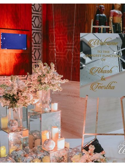 A simple yet elegant welcome sign for the Sangeet, flanked by mirrored pedestals, candles, and soft white florals, setting a glamorous tone for the evening.