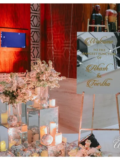 Elegant welcome signage is a must. This setup for a Sangeet function uses mirrored pedestals, candles, and floral arrangements to create a sophisticated first impression for guests.