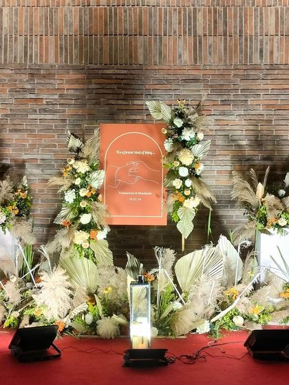 A full view of the boho-chic engagement stage. The arrangement of pampas grass, dried palms, and a custom welcome sign creates a stylish and modern backdrop against the rustic brick wall.