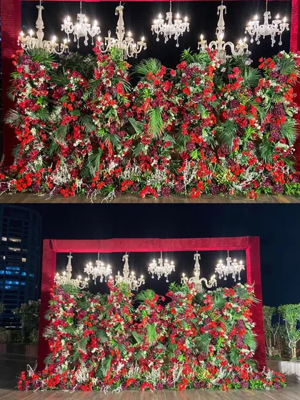 A truly dramatic reception stage featuring a dense wall of red florals and lush greenery, crowned with elegant crystal chandeliers. This design creates an atmosphere of pure opulence and romance for a grand celebration.