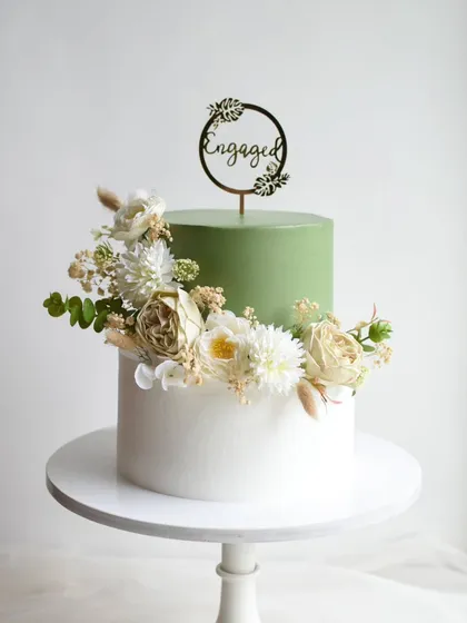 A modern and chic two-tier engagement cake in a trendy sage green and white. The design is elevated with a beautiful arrangement of fresh, natural-toned flowers for a sophisticated look.