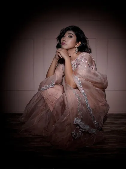 A seated portrait under a spotlight, creating an intimate and focused mood. The soft lighting beautifully highlights the delicate embroidery on the pink sharara set.
