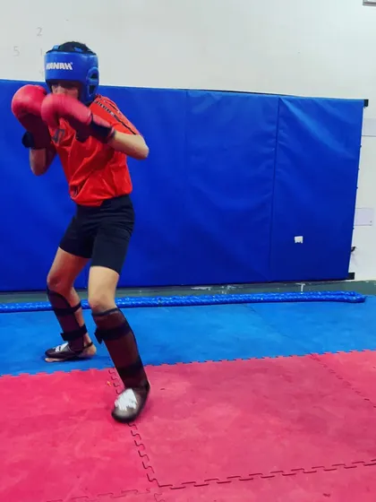 A young fighter in full sparring gear, including head guard and shin pads, maintains a solid boxing stance. This is how we prepare our students for the intensity of a real match, focusing on defense and readiness.