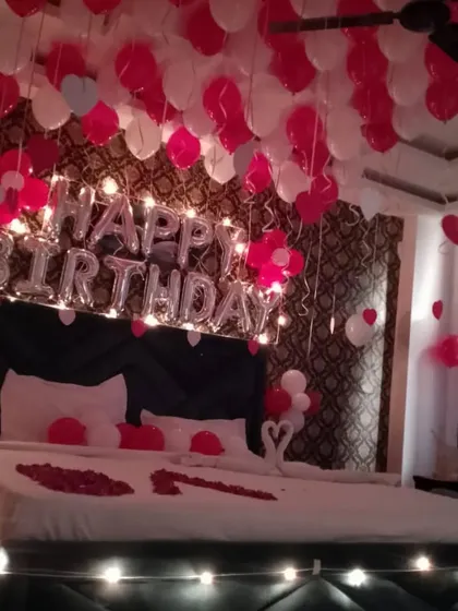 A full hotel room birthday decoration with red and white balloons on the ceiling. The bed is decorated with rose petals and towel swans for a classic romantic feel.