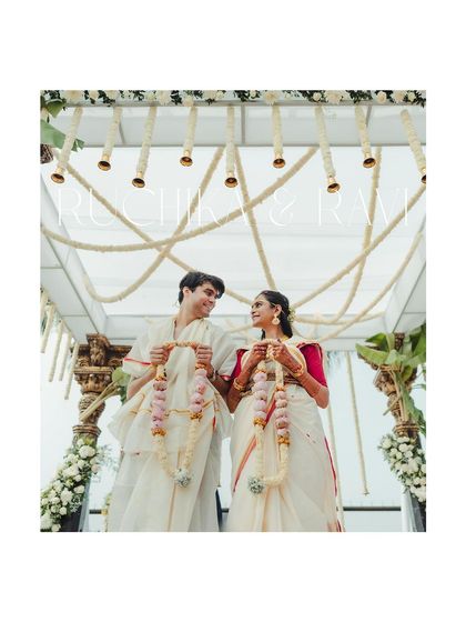 A classic portrait of the couple under their mandap. The traditional white and red attire contrasts beautifully with the floral decor, capturing the essence of a South Indian wedding.