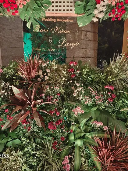 A lush, tropical-themed photo booth for a wedding reception, featuring dense greenery and flowers with a personalized sign for the couple.