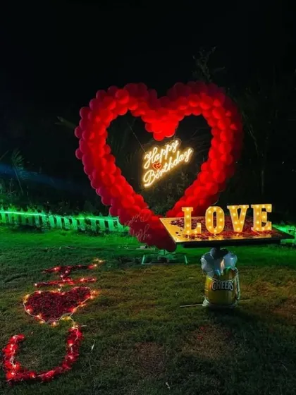 A grand romantic gesture in a garden at night. A large heart-shaped balloon arch surrounds a 'Happy Birthday' neon sign, with 'LOVE' marquee letters and a path of rose petals.