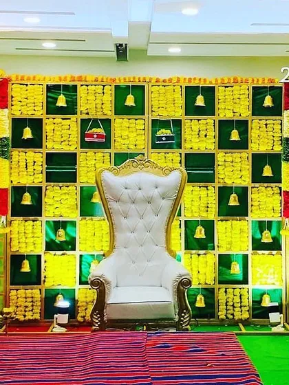 A classic baby shower backdrop with a checkerboard pattern of woven leaves and marigolds. The hanging bells and a traditional striped mat on the floor add to the authentic and festive feel.