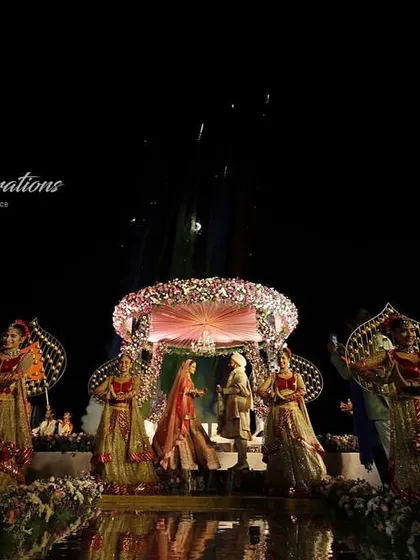 A stunning wide shot of a Jaimala ceremony on a stage set over the water at a Goa wedding. The reflection, fireworks, and dancers create a breathtakingly beautiful and unique memory.