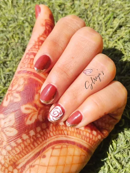 A beautiful combination of deep maroon gel polish with a delicate, hand-painted white rose on the ring finger. The subtle gold glitter on the cuticles adds a touch of festive sparkle that pairs perfectly with henna.