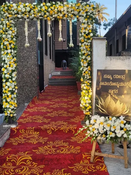 I create welcoming entrances that set the tone for the event. This one features a grand floral arch of white and yellow flowers over a red carpet, with a custom welcome sign in Kannada to greet the guests.