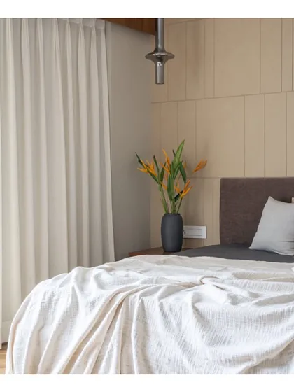 This bedroom in 'Vishu Home' is a study in calm neutrality. The beige paneled wall, dark wood headboard, and soft white linens come together to create a peaceful and sophisticated space for rest.
