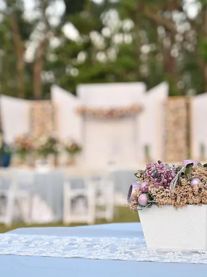 A simple yet elegant centerpiece with dried lavender and pastel flowers on a guest table, with the outdoor wedding stage blurred in the background.