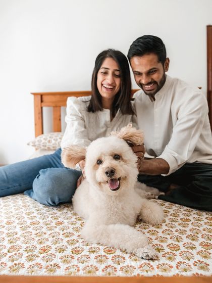A happy family moment on the bed. Dinkan, a fluffy white dog, is the center of attention, clearly enjoying the love from his smiling parents.