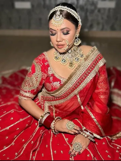 A bride seated, showcasing the full spread of her red lehenga with silver embroidery. The look is completed with a pearl-bordered sheesh patti.
