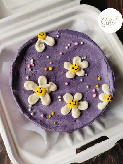 A happy and vibrant purple bento cake. The design is dotted with smiley-faced flowers and colorful sprinkles, making it a cheerful treat for any occasion.