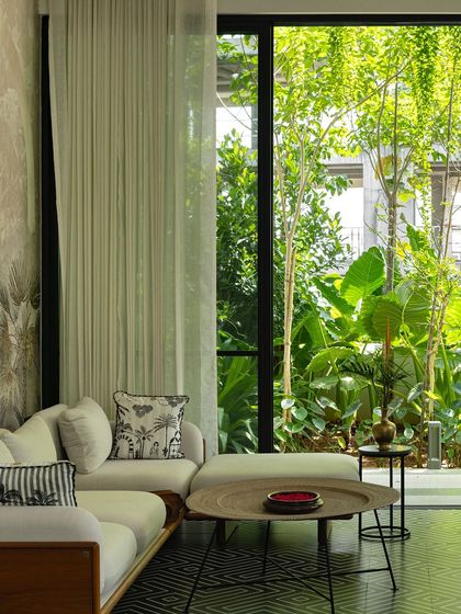A bright, airy living room with large windows that open up to a view of a lush, tropical garden. The interior decor, with its natural tones and textures, complements the green vista, creating a cohesive and calming atmosphere.