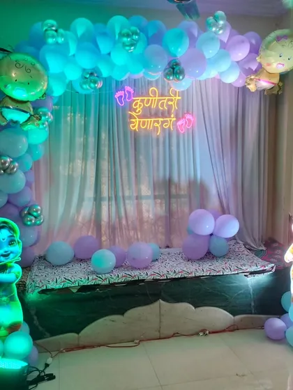 A baby shower decoration for a smaller space, featuring a balloon arch over a seating area, a neon sign, and illuminated Radha Krishna cutouts.