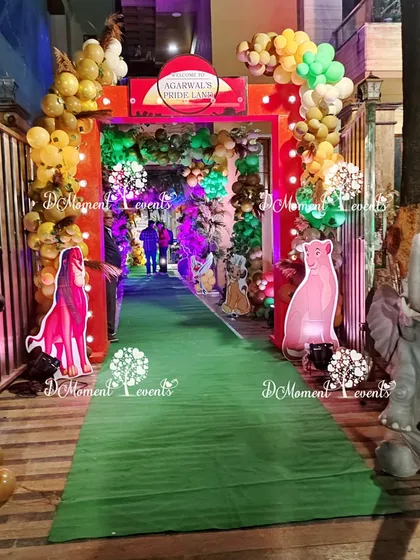 A grand entrance to a Lion King themed baby welcome party. A red archway and a green carpet path are lined with 3D animal props like a zebra and elephant, creating an exciting safari welcome for guests.