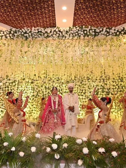 An epic bride and groom Jaimala on a stage adorned with a cascade of white flowers. Dancers in traditional attire perform around them, adding a layer of grace and celebration to the ceremony.