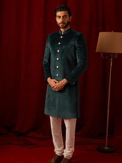 A full-length view of the deep green velvet bandhgala, paired with classic white trousers. The simplicity of the styling allows the color and fabric of the jacket to truly shine.