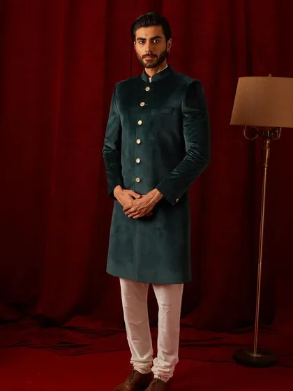 A full-length view of the deep green velvet bandhgala, paired with classic white trousers. The simplicity of the styling allows the color and fabric of the jacket to truly shine.