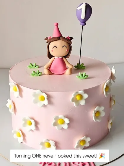 A sweet first birthday cake with a simple and charming design. The pink cake is decorated with daisies and a cute fondant girl topper holding a balloon.