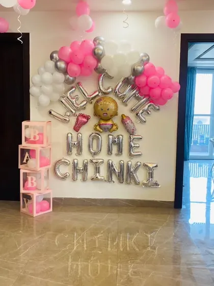 A personalized "Welcome Home Chinki" decoration to make the moment unforgettable. This setup uses silver foil letter balloons, pink 'BABY' blocks, and clusters of pink and silver balloons to create a loving atmosphere.