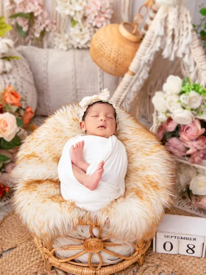A newborn wrapped in classic white rests in a boho-style chair, showcasing a simple yet elegant setup with natural textures and soft florals. This highlights our versatile approach to newborn photography themes.