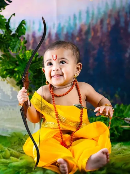 A joyful little boy dressed as Sri Rama, holding his bow and arrow. The forest backdrop is perfect for recreating scenes from the Ramayana.