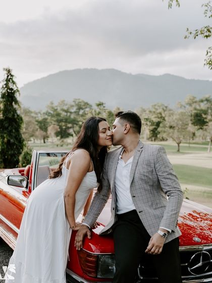 An intimate close-up of a couple sharing a kiss against the hood of a vintage car in Thailand. This romantic shot focuses on their connection, with the beautiful natural scenery blurred in the background.