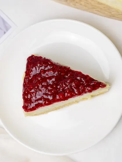 A slice of cheesecake with a vibrant strawberry or raspberry topping. The minimalist presentation on a white plate highlights the simple elegance of the dessert.