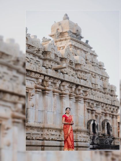 A wide shot of the bride in her red saree standing before the magnificent temple facade, emphasizing the grandeur of the wedding venue.