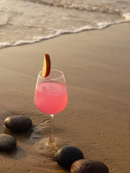 A beautiful pink cocktail in a wine glass, set against the sand and waves at sunset. The soft light and natural elements create a very romantic and dreamy mood.