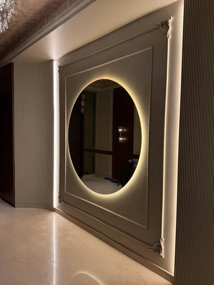 A stunning entrance foyer featuring a large, round mirror with warm backlighting. The design uses wall molding and vertical strip lights to create a grand and elegant first impression.
