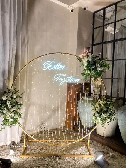Simple, modern, and elegant. This engagement decor features a large golden ring with a "Better Together" neon sign and cascading fairy lights, flanked by arrangements of classic white flowers.