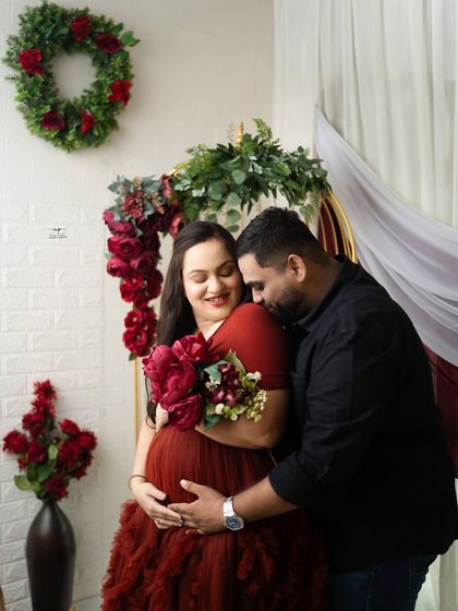 An intimate portrait of an expecting couple, framed by a festive wreath and red flowers. The husband's gentle embrace from behind shows their deep connection and shared joy.