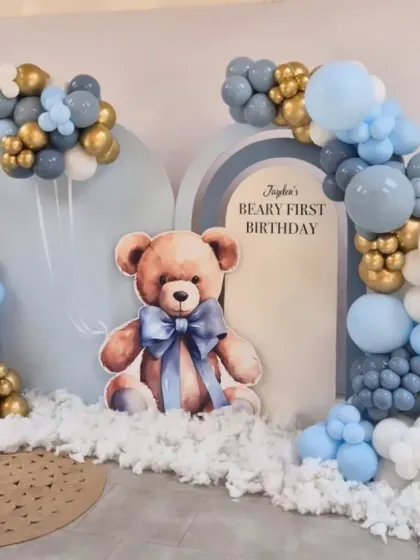 A "Beary First Birthday" theme with a large teddy bear cutout, arched backdrops, and a soft blue, grey, and gold balloon arrangement.