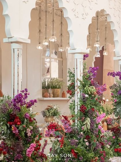 A lush and floral-rich lounge area, where towering arrangements of purple and red flowers are interspersed between comfortable seating groups against a backdrop of white arches.