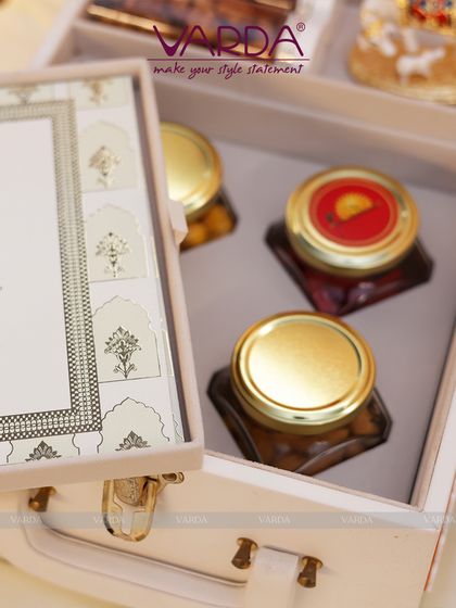 A close-up of a white wooden keepsake box designed like a small suitcase. The interior holds gourmet jars and a framed invitation, showcasing a blend of traditional Indian patterns with a clean, modern aesthetic.